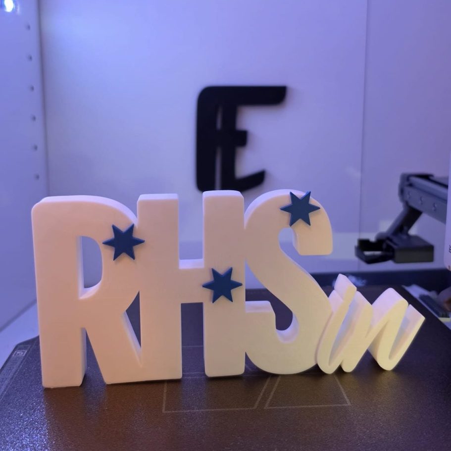Bunte Buchstaben "RHS" mit blauen Sternen auf einem Tisch hinter einer undeutlichen Fläche.